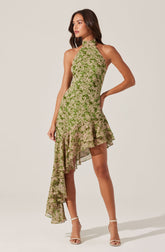 A woman is wearing a sleeveless, high-neck dress with a green and beige floral pattern. The dress features an asymmetrical hemline, with a shorter hem on one side and a longer, ruffled section cascading down to the floor. She is also wearing beige high-heeled sandals with ankle straps.